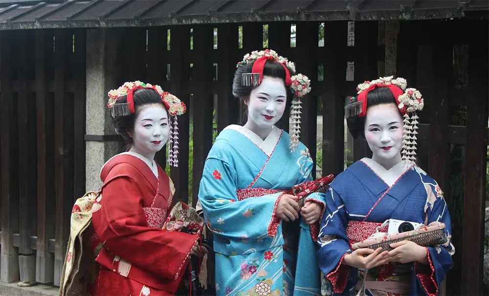 Three geisha in traditional kimonos smiling outdoors.