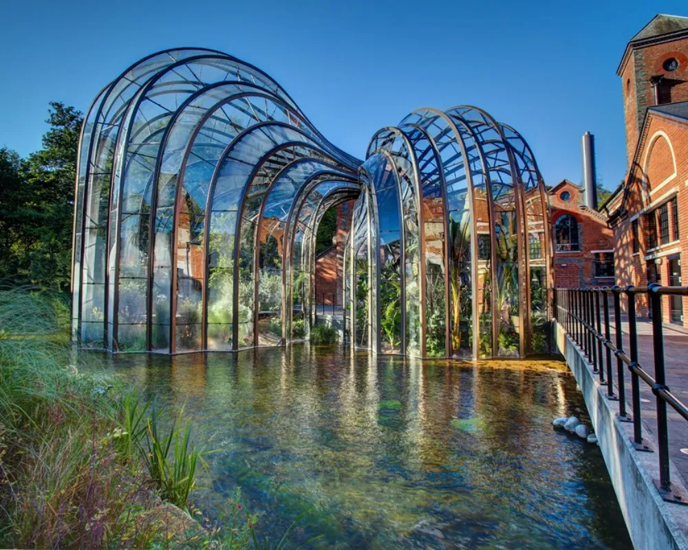 Architectural glasshouse structure by pond with industrial backdrop.