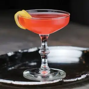 Red cocktail in a coupe glass with lemon twist.