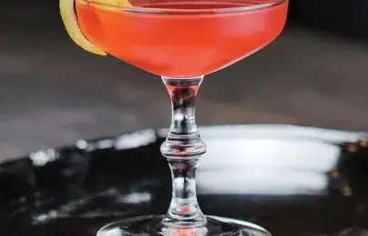 Red cocktail in a coupe glass with lemon twist.